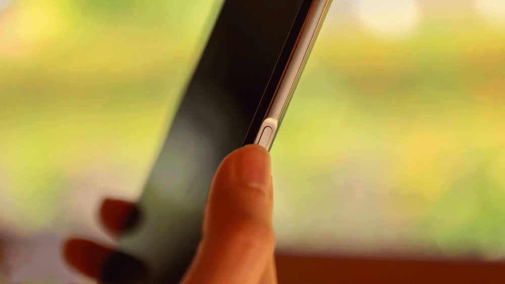 Main tenant un smartphone à l'écran éteint, le pouce posé sur le bouton d'alimentation